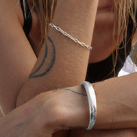 Close-up of a wrist adorned with a sleek silver cuff bracelet and a delicate silver chain bracelet. The arm features a bold crescent moon tattoo, and the background showcases a casual outdoor vibe.