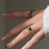 Person's hand with two rings: a silver ring with a double drop design on the index finger and a gold ring with a wavy shape on the ring finger. The nails have minimalist black dot designs.