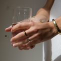 a hand holding a glass of water and wearing gold jewelry. A wavy-shaped gold ring is on the middle finger, a simple bamboo design gold band is on the ring finger, and a gold cuff bracelet is around the wrist. Little black dots are used to decorate the nails.