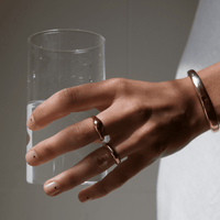 a hand holding a glass of water and wearing gold jewelry. A wavy-shaped gold ring is on the middle finger, a simple gold band is on the ring finger, and a gold cuff bracelet is around the wrist. Little black dots are used to decorate the nails.