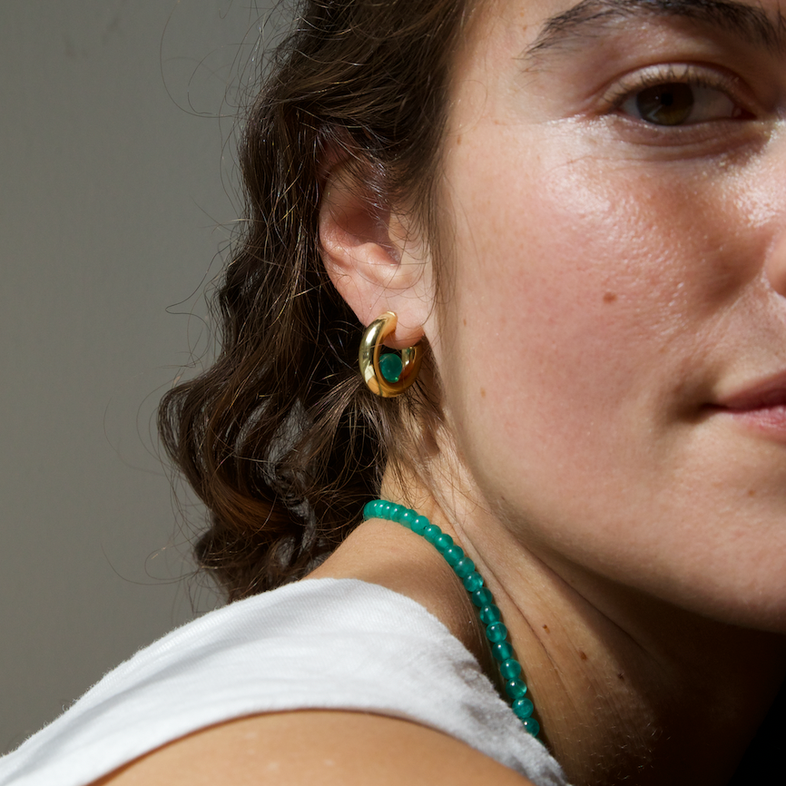a woman with curly hair, wearing a gold hoop earring with a green quartz gemstone and a green quartz necklace