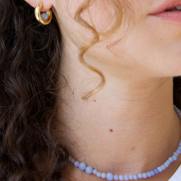 a woman with curly hair wearing a gold hoop earring with a blue lace agate gemstone. She also wears a blue lace agate gemstone necklace