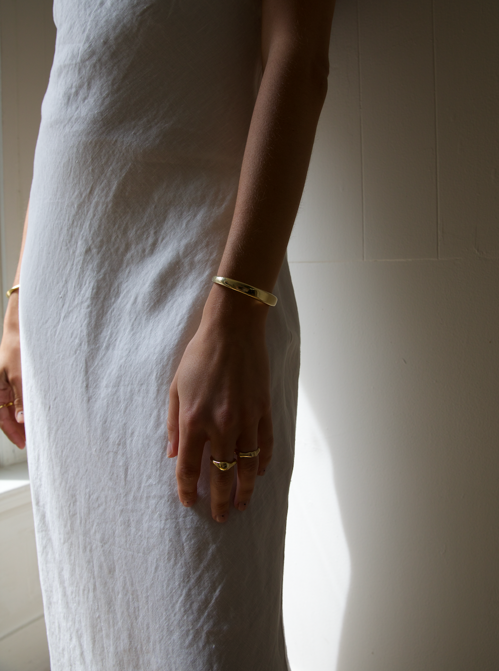 Close-up of a woman’s hand and wrist, wearing a gold cuff bracelet and rings, against a white dress. The simple and elegant jewelry complements the minimalist style of her outfit.