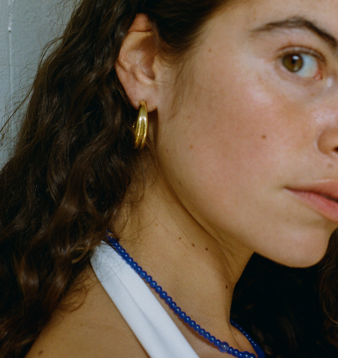 Close-up portrait of a woman wearing a blue beaded necklace and a large gold hoop earring. Her wavy brown hair falls loosely, and she wears a white top. 