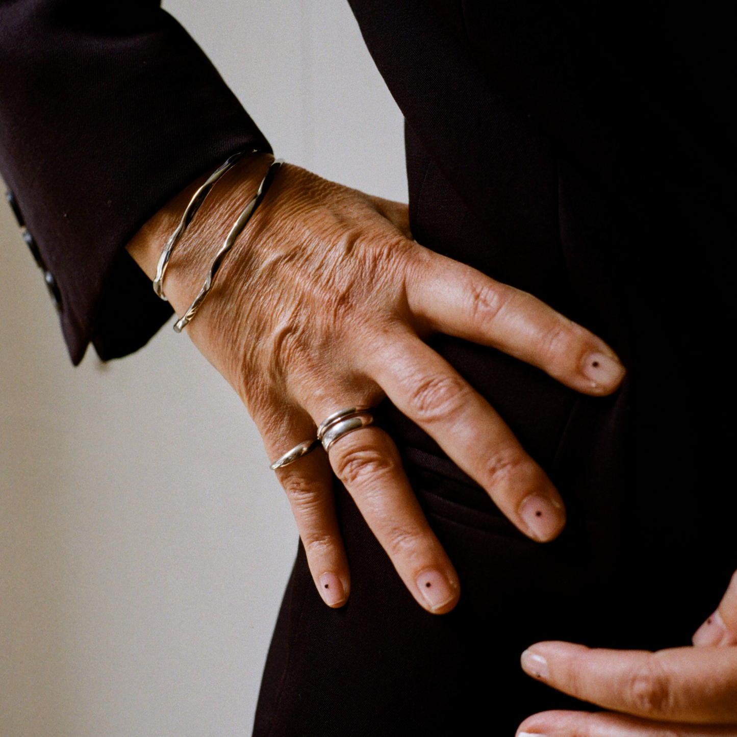 A hand with a silver ring on the ring finger and a silver ring on the pinkie. The wrist has two uneven silver cuff bands. The hand is sitting on the hip, and there are small black dots on the nails.