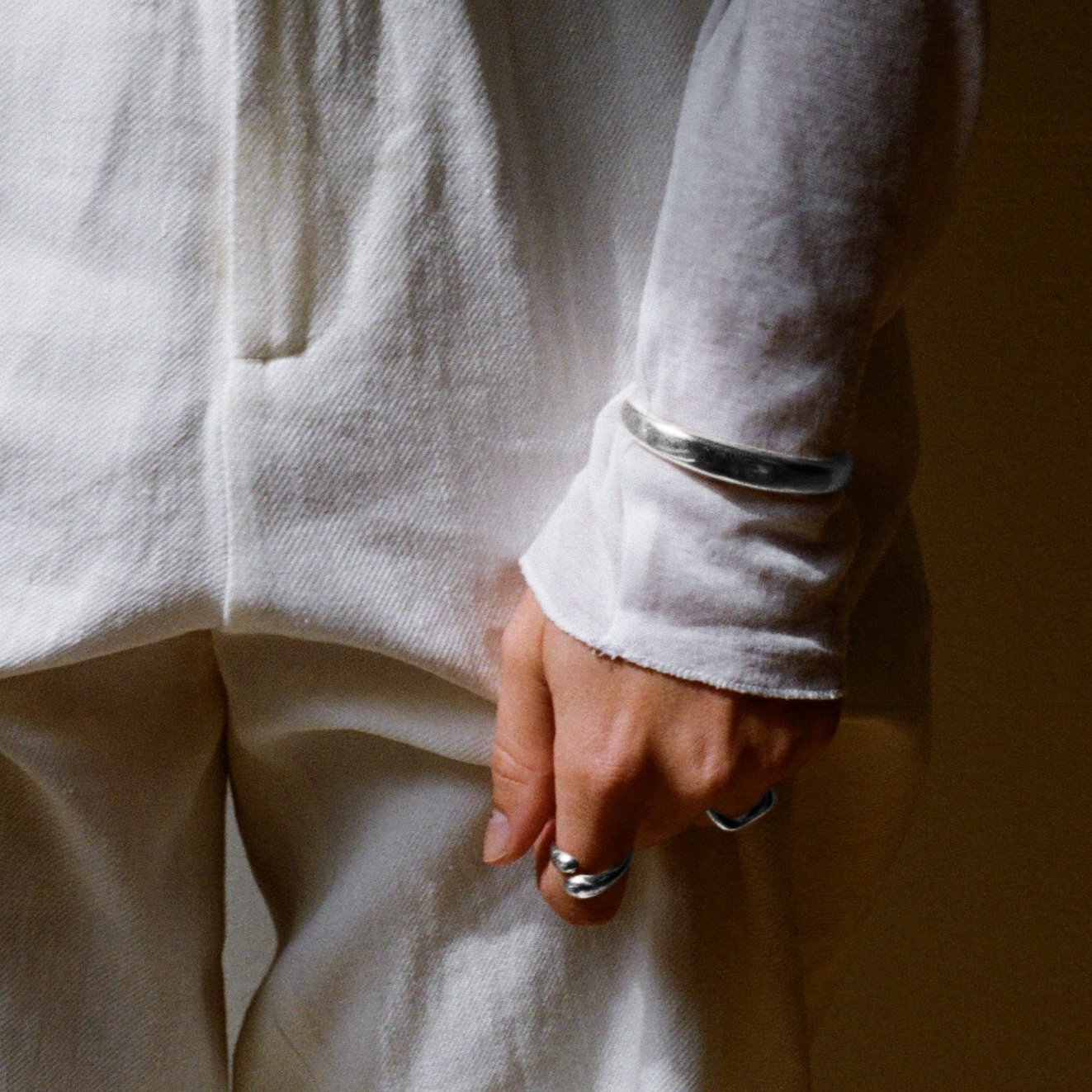 Close-up of a hand wearing a silver cuff bracelet and two sculptural silver rings, styled with an oversized white shirt and matching pants. The minimalist jewelry adds a sleek, modern touch to the monochromatic outfit.