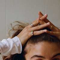 Hands with different rings on them rest on top of someone's head. On one hand, the pinkie has a silver ring and the middle finger has a double band ring. On the other hand, the ring finger has a gold ring. The person is dressed in a white long-sleeved shirt and has curly hair.