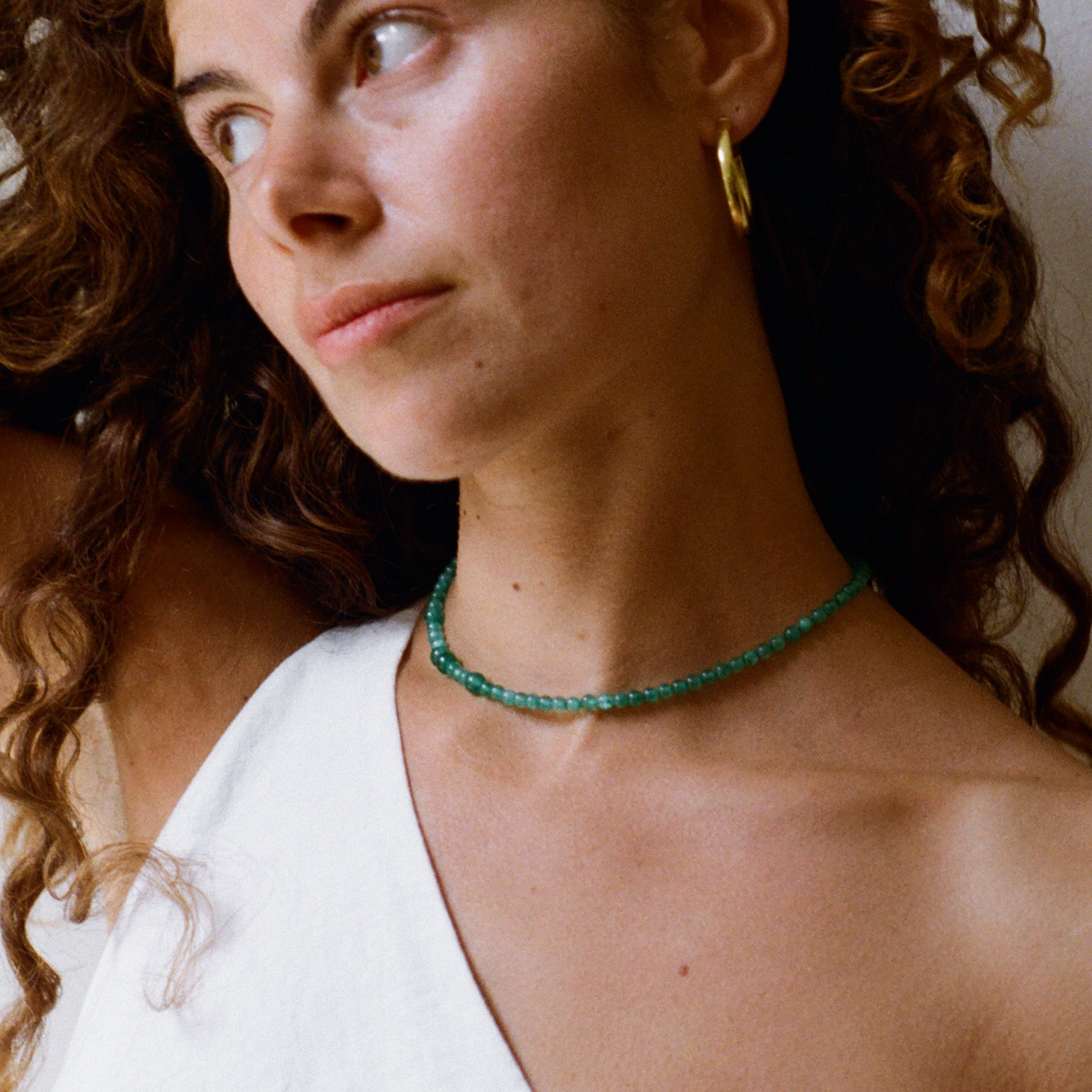 Curly hair woman with a one shoulder shirt looking outside, wearing a green quartz gemstone necklace and a bold gold hoop