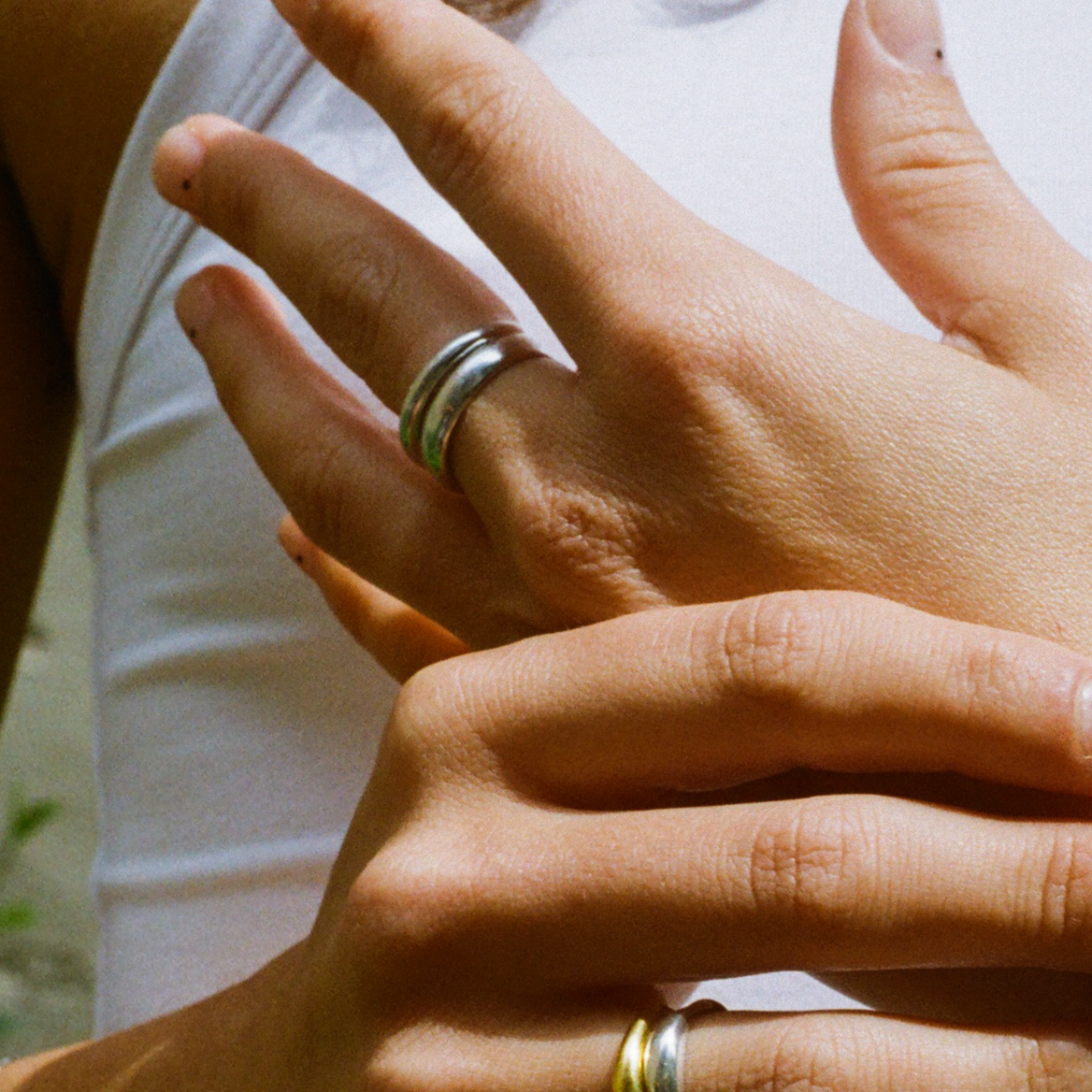 A close-up of a person's hands, showing a silver double-banded ring on the middle finger and a gold ring on the ring finger of one hand. The person is wearing a white sleeveless top and has minimalistic nail art with small black dots