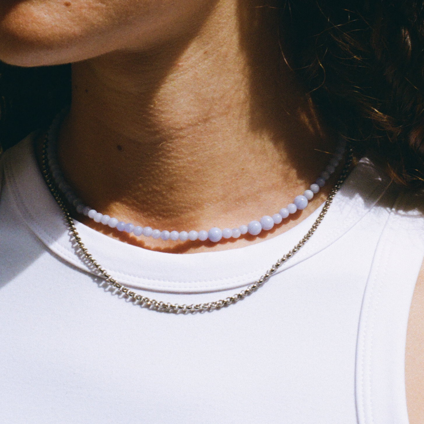 Person wearing a white top, with a blue lace agate gemstone necklace and a silver rolo chain necklace