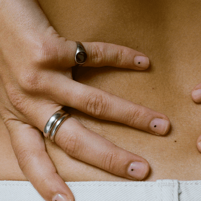 Hand with silver rings on the middle and ring fingers. The middle finger has a ring with double bands, and the ring finger has a round signet ring. There are small black dots on the nails.