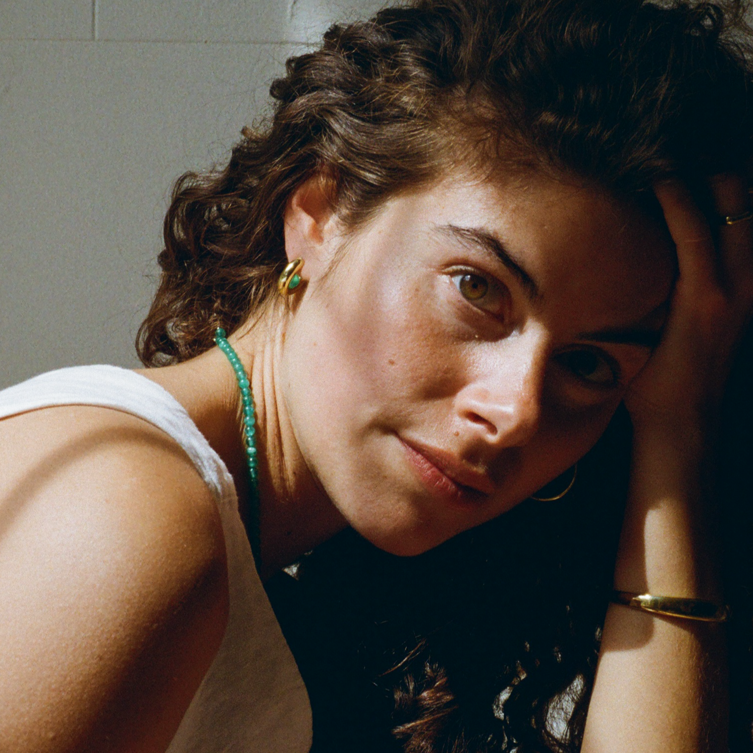 woman with curly hair, wearing a gold hoop earring with a green quartz gemstone, a green quartz gemstone necklace, and a golden bold cuff bracelet.