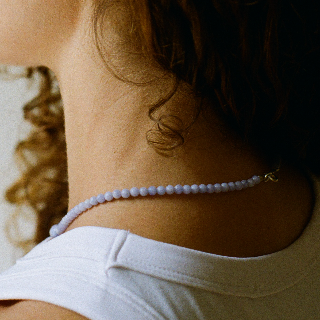 Back of a curly hair woman, wearing a white top, showing the back of a blue lace agate gemstone necklace.