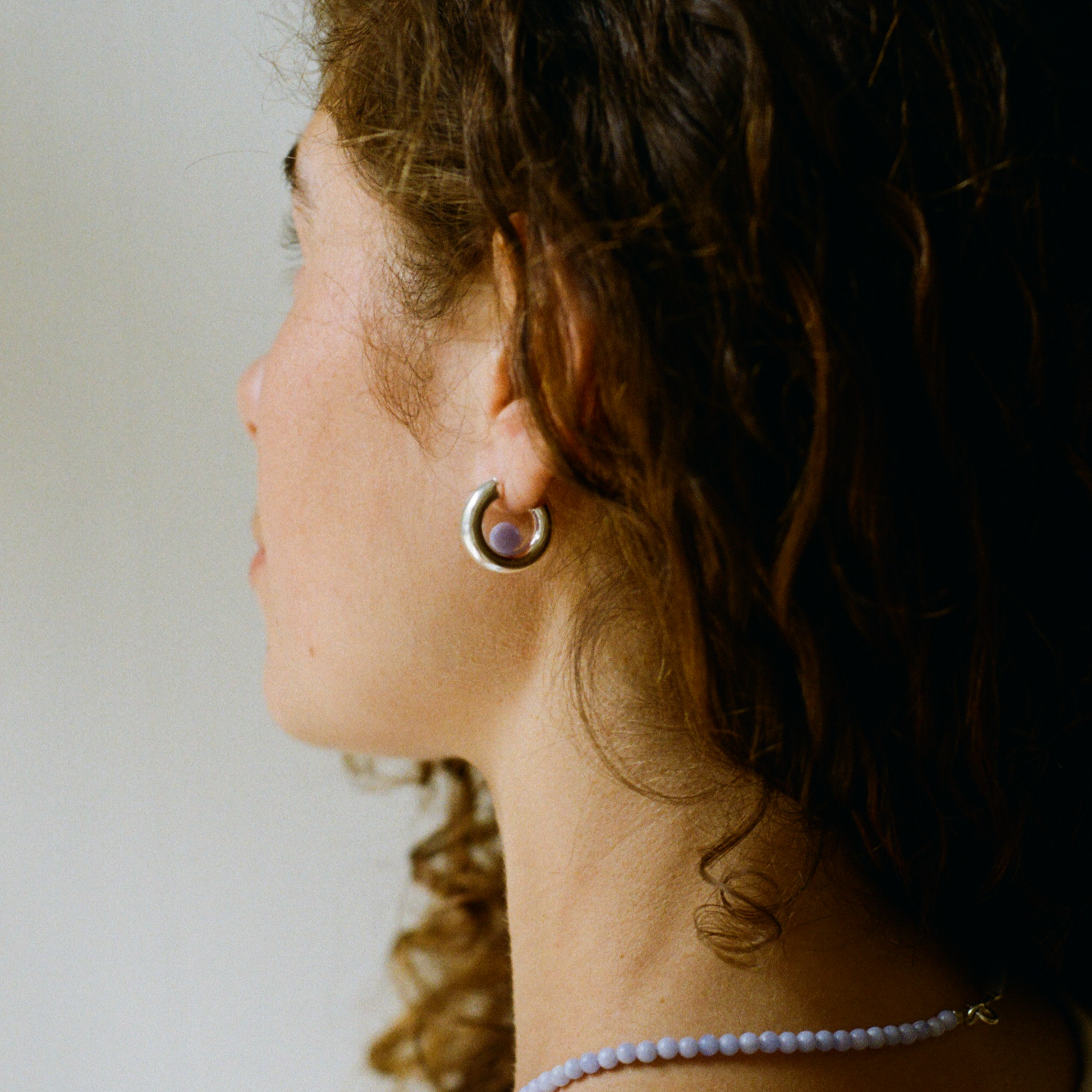 side profile of a woman with curly hair wearing a silver hoop earring with a blue lace agate gemstone. She also wears a blue lace agate gemstone necklace