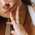 A person with a gold uneven bold ring on their finger with their hand on their chin. They are wearing a blue beaded necklace