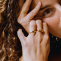 Person with their hand on their face with gold jewelry. The middle finger is wearing a wavy gold ring, the ring finger has a bamboo gold ring, and the other hand with a triple line ring.