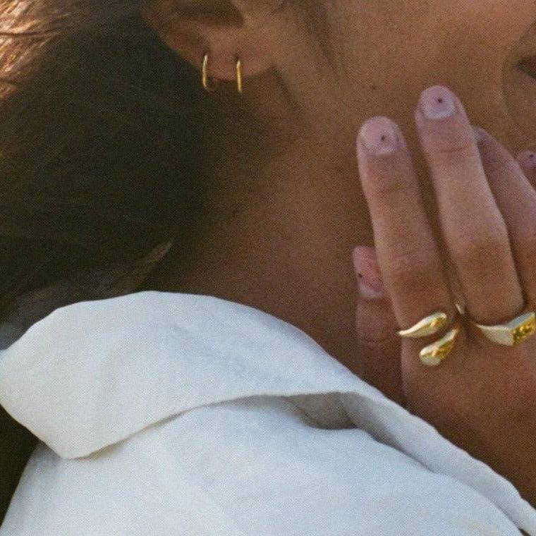 Close-up of a model wearing small gold hoop earrings and two sculptural gold rings with a bold design, styled with a white collared shirt. The jewelry adds a modern and elegant touch.