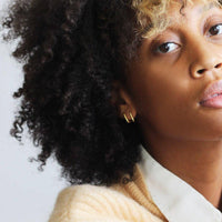 Close-up of a model wearing small gold hoop earrings and two sculptural gold rings with a bold design, styled with a white collared shirt. The jewelry adds a modern and elegant touch.